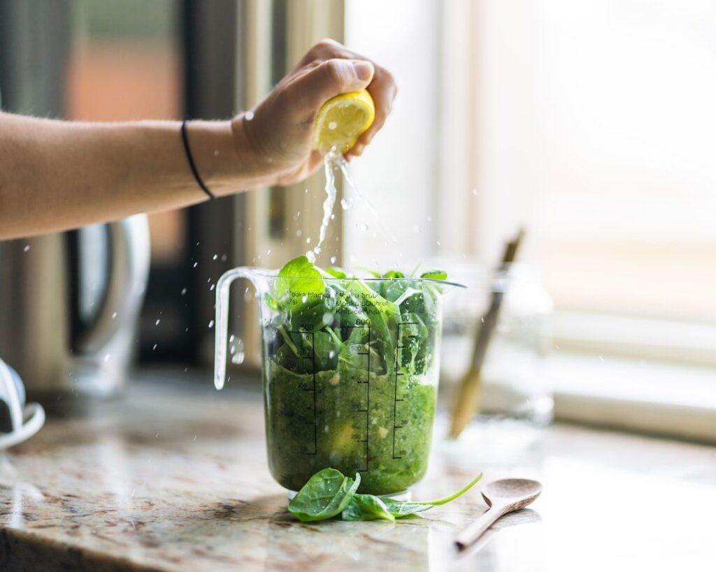 En hand pressar färsk citron över en grön smoothie med spenat i en måttbägare.